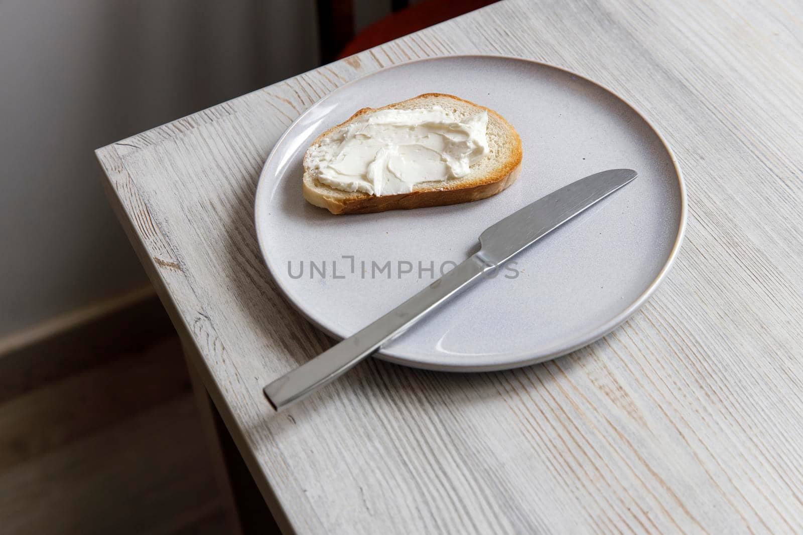 A piece of bread with curd cheese spread on it on a white ceramic plate with a knife on the beige table. by elenarostunova