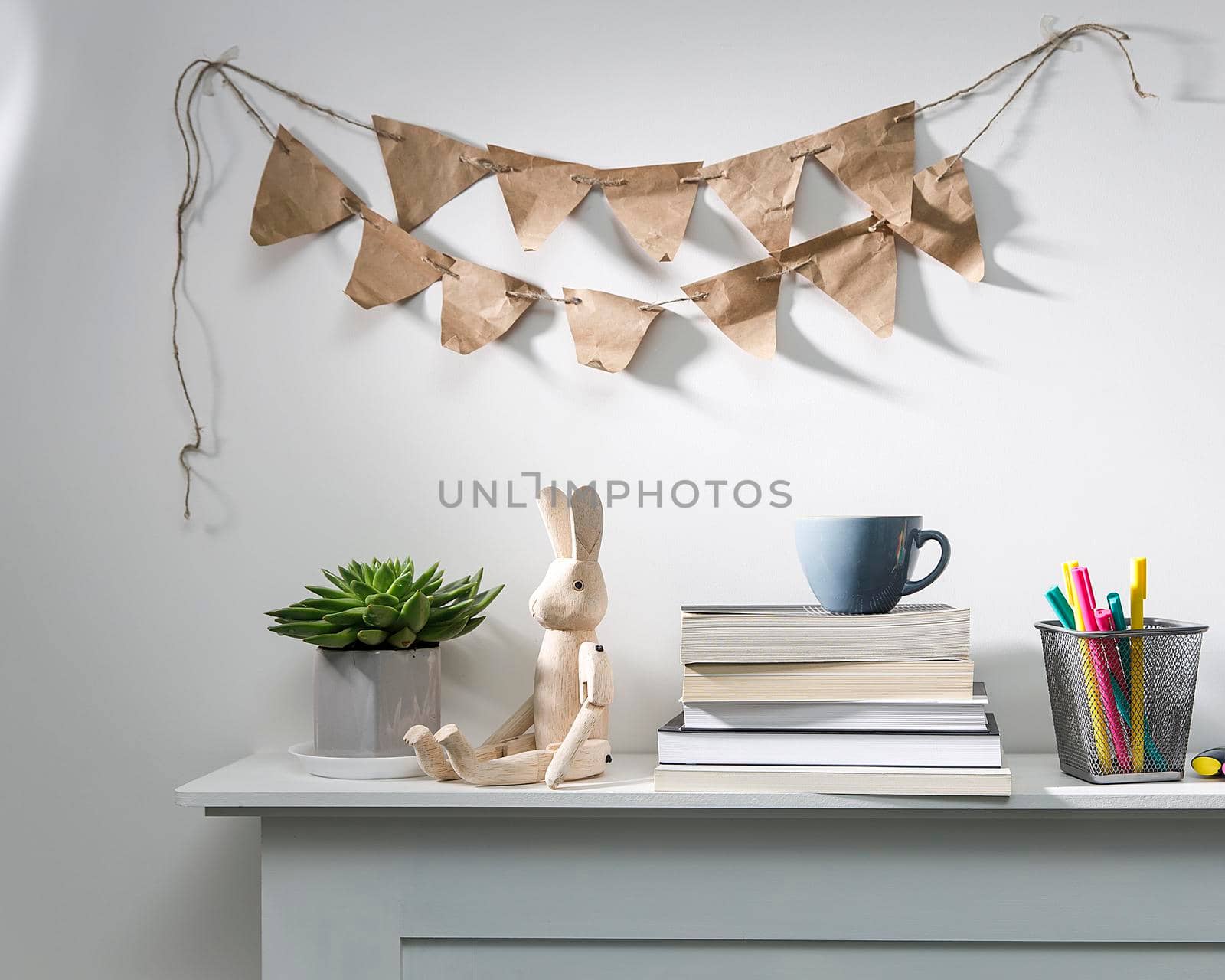 Echeveria in geometric pot, stack of books, pens in a pencil holder, felt-tip pens, wooden figurine of a hare are on shelf. Garland of craft paper flags on the wall above the shelf. Place for text by elenarostunova