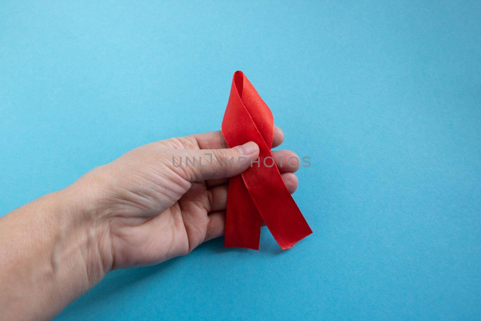 A hand holds a red ribbon on a blue background - the concept of World AIDS Day, Donor Day, Hemophilia Day.