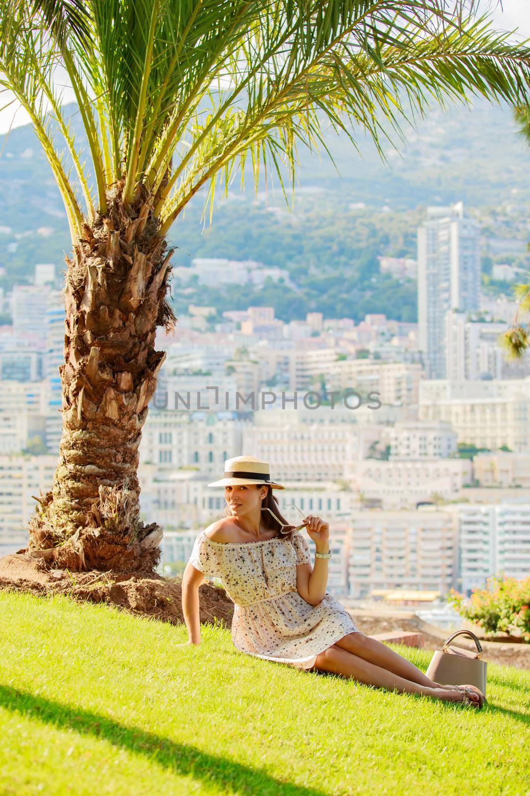 a bright beautiful girl in a light dress and hat sits on the grass under a palm tree in Monaco in sunny weather in summer, streets of old town of Monaco. High quality photo