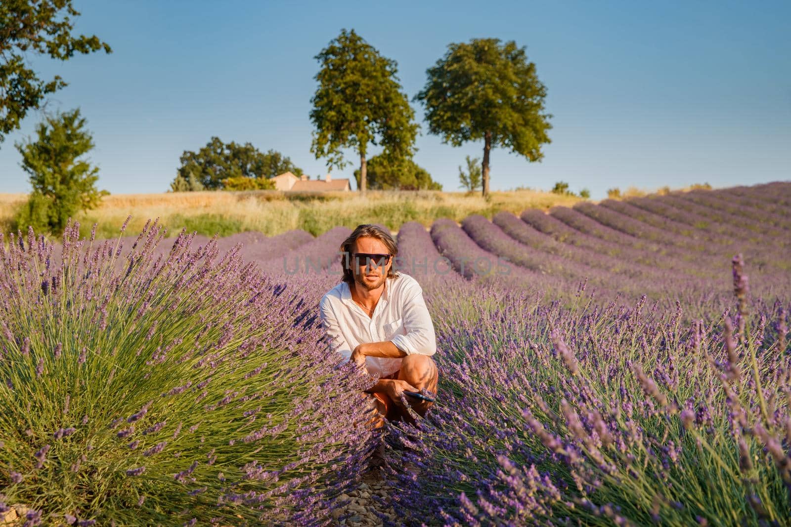 The handsome brutal man with long brunette hair sits on field of lavender in provence near Valensole, France, clear sunny weather, in a rows of lavender, red shorts, white shirts, blue sky by vladimirdrozdin