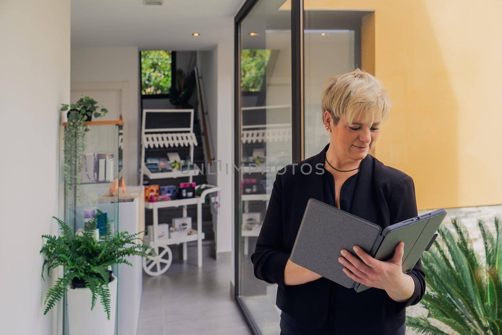 Smiling mature professional beautician with blonde hair, dressed in black work uniform, checking email on her Tablet. owner in her small beauty salon looking at laptop. Work from home, enterprising woman, active small business owner. Relaxed atmosphere and soft lighting from window, natural light, table, computer. Horizontal