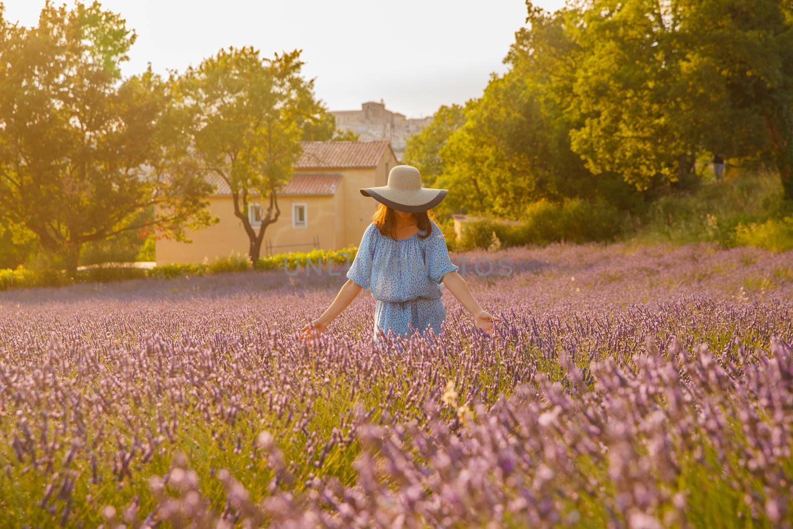 The beautiful young girl in a blue dress and cap walks across the field of a lavender, long curly hair, smile, pleasure, a house of the gardener in the background, trees, perspective of a lavender. High quality photo