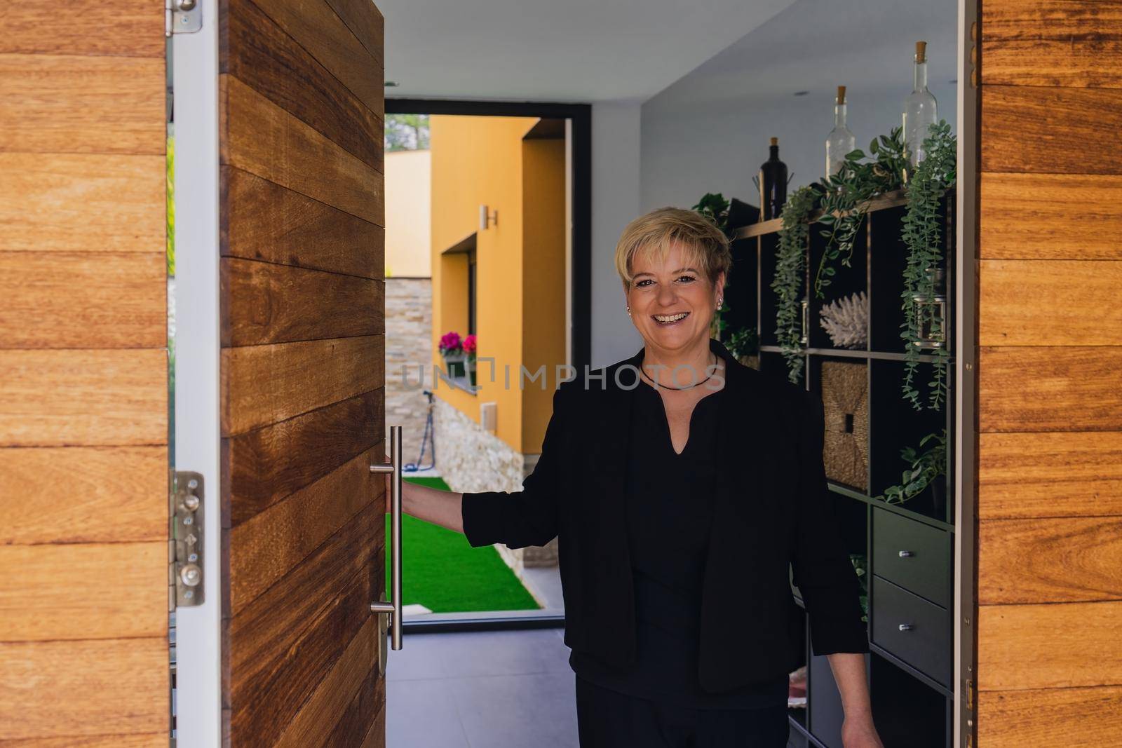 Smiling mature professional beautician woman with blonde hair, dressed in black work uniform, opening the door for visitors. . Work from home, freelance, business, creative occupation, lifestyle concept.Relaxed atmosphere and soft lighting from interior windows, natural light from outside, entrance door. Horizontal.