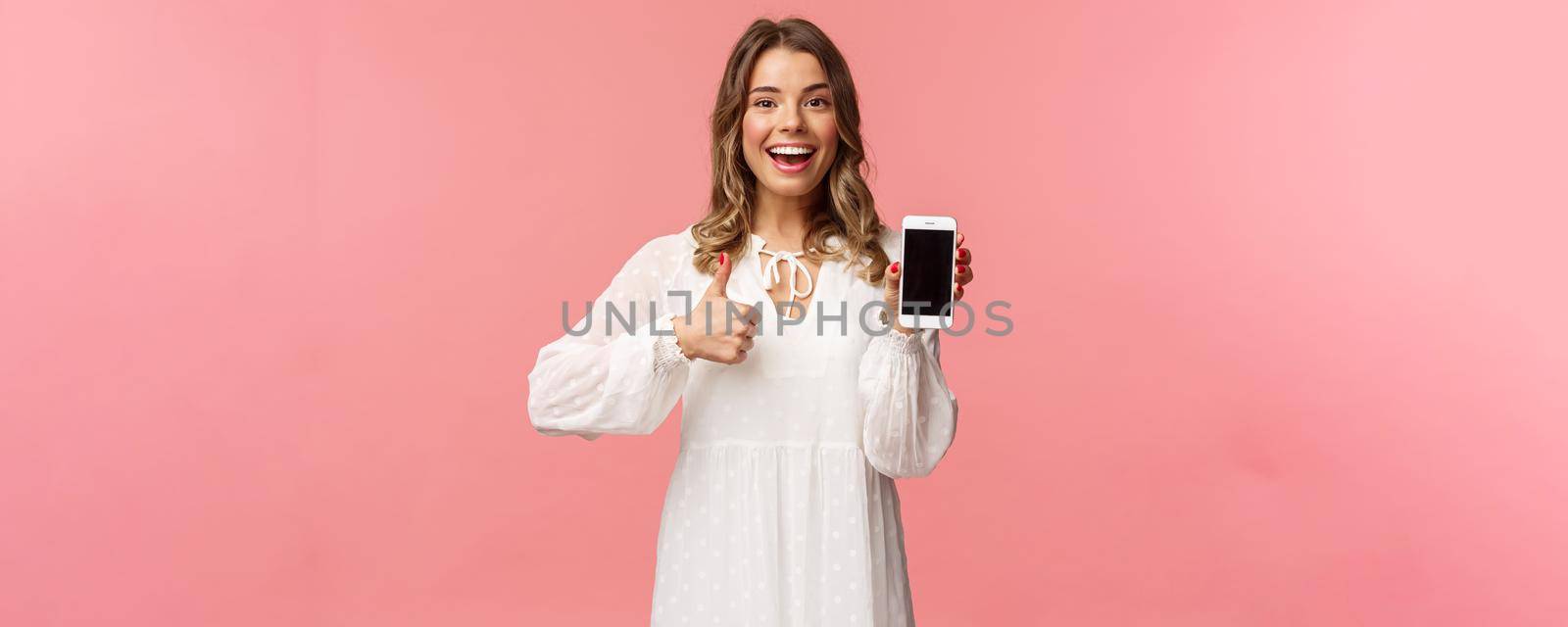 Portrait of upbeat good-looking blond woman in white dress, recommend download app or subscribe, using online shopping application, make thumb-up, show mobile phone screen.