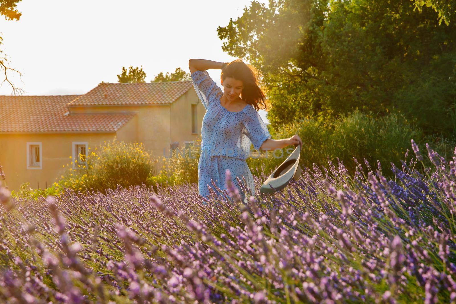 The beautiful young girl in a blue dress and cap walks across the field of a lavender, long curly hair, smile, pleasure, a house of the gardener in the background, trees, perspective of a lavender. High quality photo