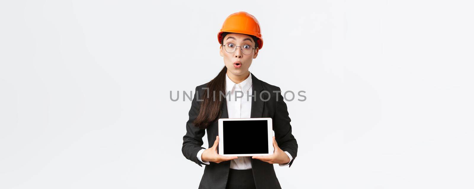 Impressed asian female construction engineer, businesswoman in safety helmet and suit showing chart, introduce digram at digial tablet screen, standing amazed over white background.