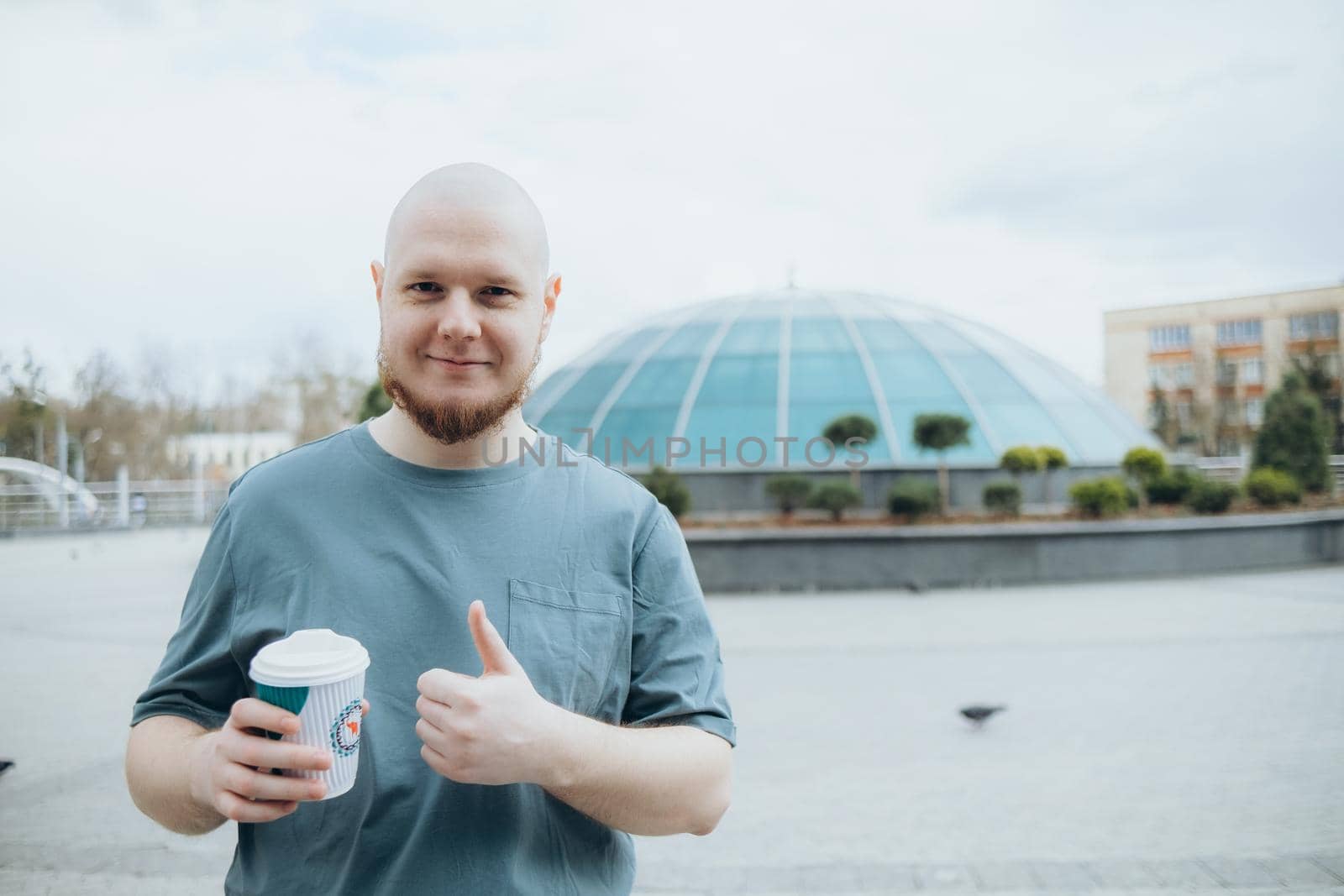 Bald bearded man drinking coffee outdoors and smiling thumb up