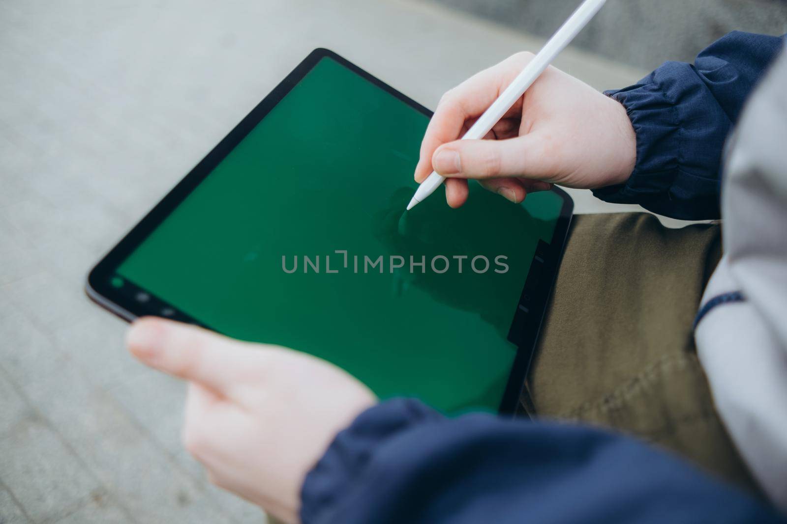 From below of hipster man using modern tablet with stylus while creating new project and sitting on street near glass skyscraper