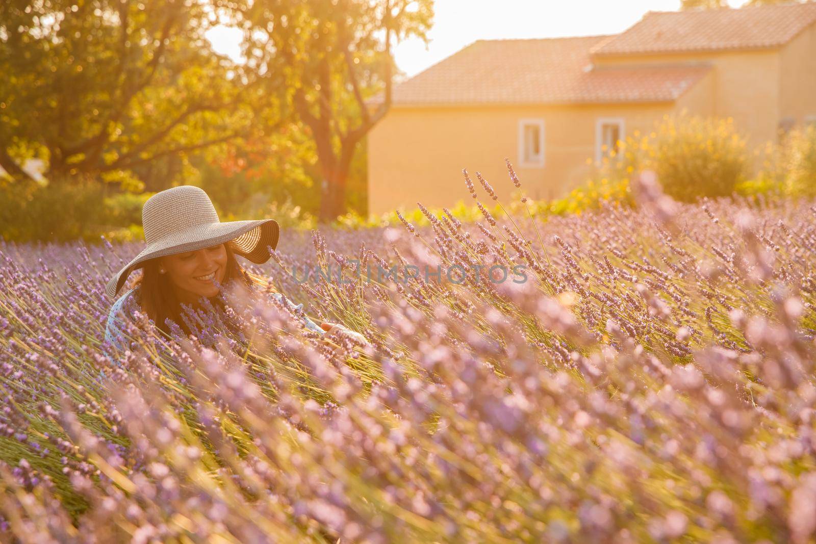 The beautiful young girl in a blue dress and cap sits in a lavender flowers, long curly hair, smile, pleasure, a house of the gardener in the background, trees, perspective of a lavender by vladimirdrozdin