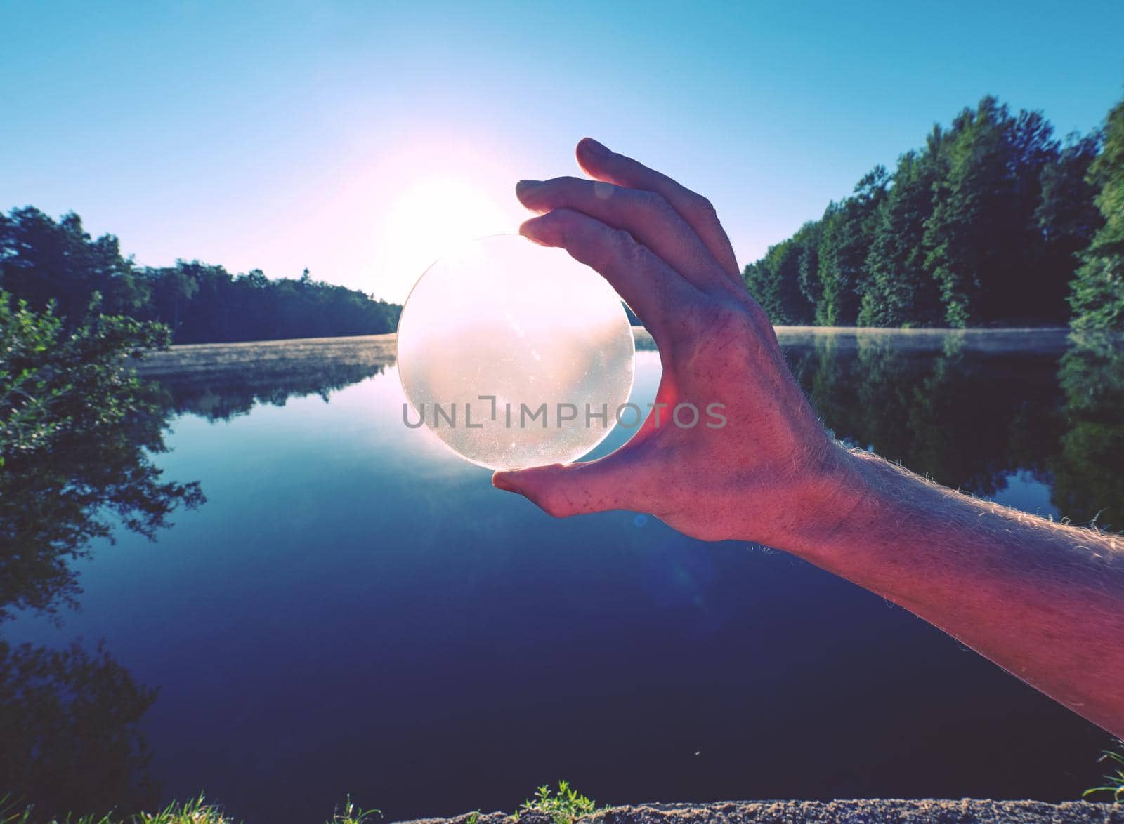 Looking through glass lens. Hand hold glass ball withuniquely reflects  summer lake scene with sunset.