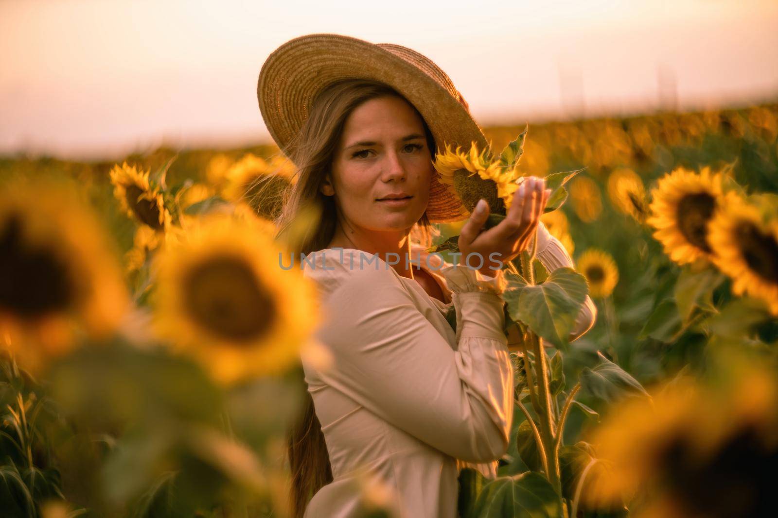 Beautiful middle aged woman looks good in a hat enjoying nature in a field of sunflowers at sunset. Summer. Attractive brunette with long healthy hair. by Matiunina