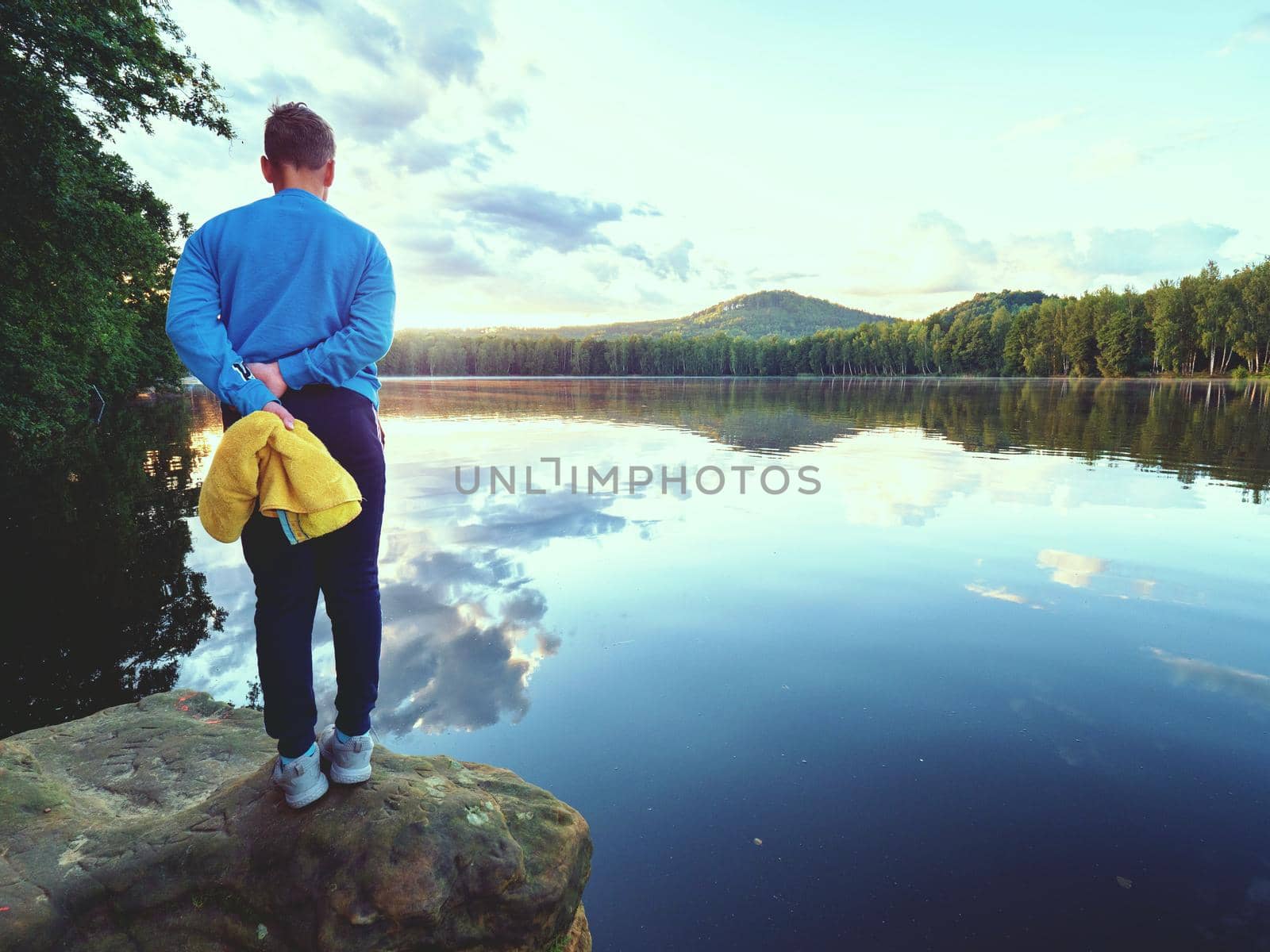 Young boy with yellow towel and swimming goggles in hands stay in morning on jumping stone above lake water. Natural summer holiday resort.