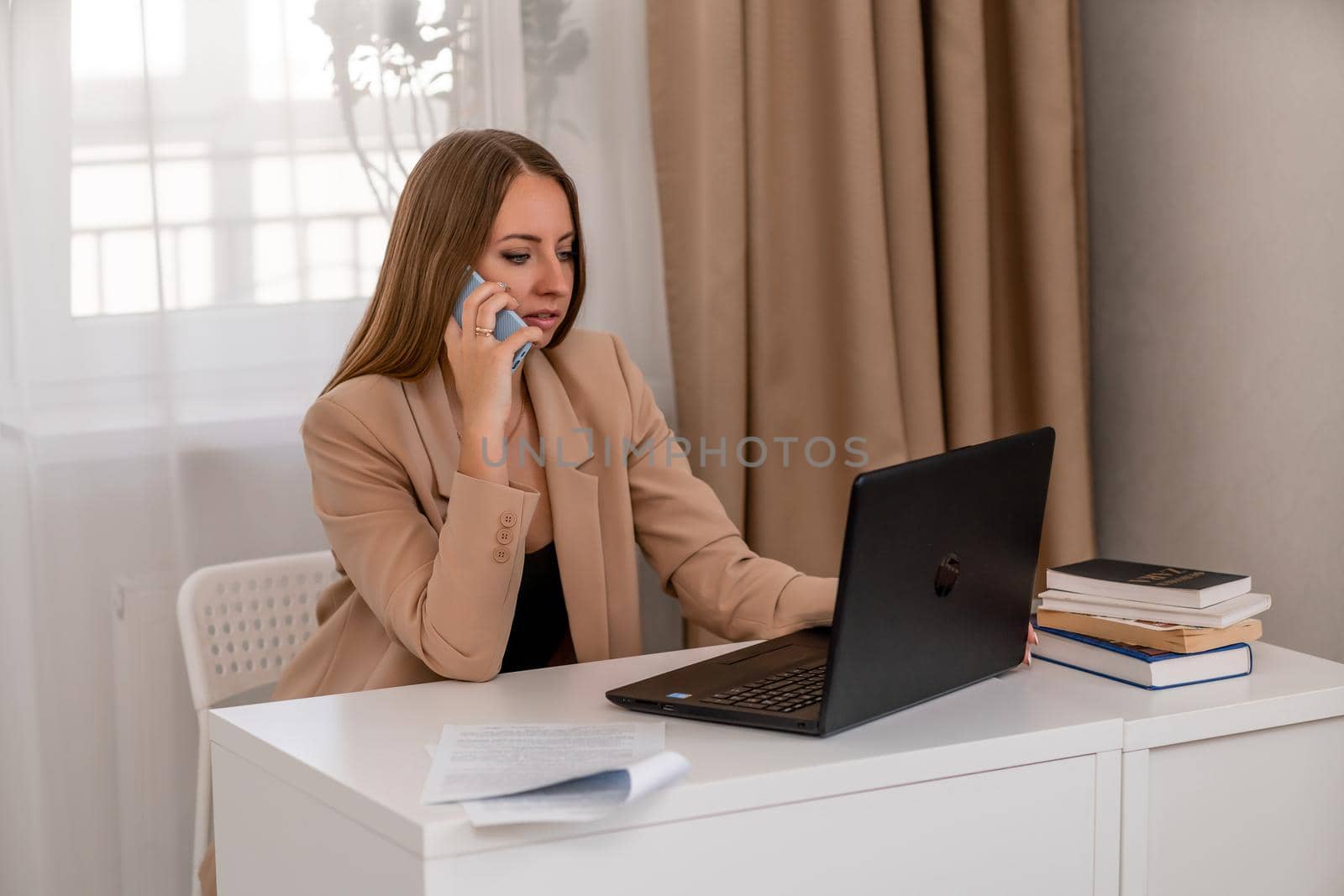 European professional woman is sitting with a laptop at a table in a home office, a positive woman is studying while working on a PC. She is wearing a beige jacket and jeans and is on the phone