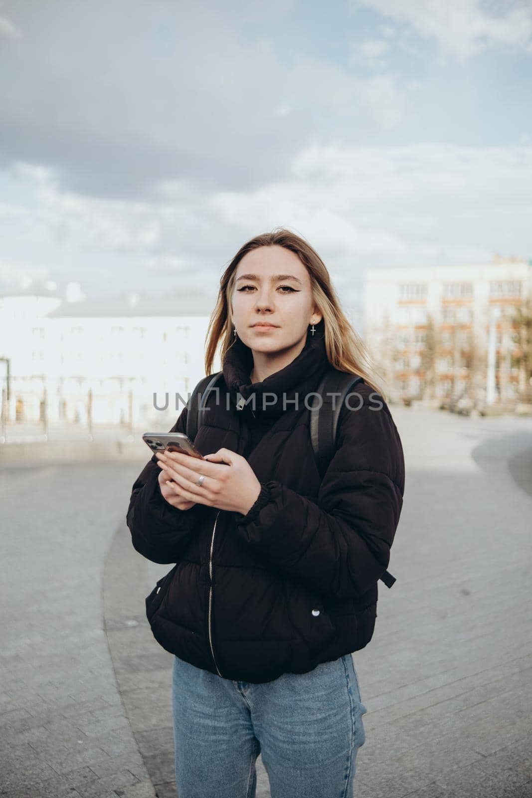 gorgeous beautiful young woman with blonde hair messaging on the smart-phone at the city street background. pretty girl having smart phone conversation in sun flare.