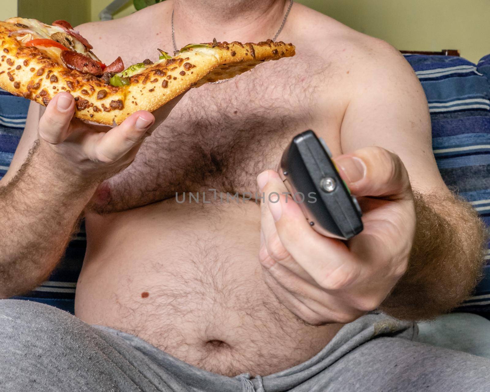 Fat man eating pizza and using TV remote, unhealthy lifestyle concept