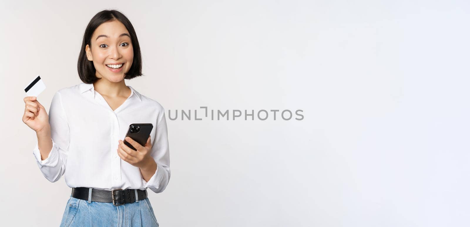 Online shopping concept. Image of young asian modern woman holding credit card and smartphone, buying with smartphone app, paying contactless, standing over white background.