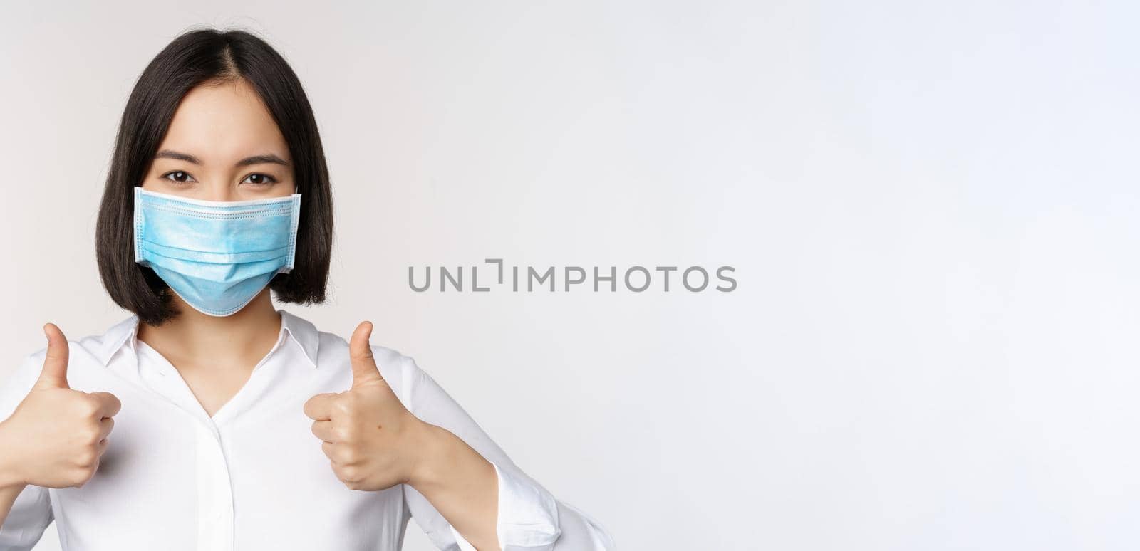 Portrait of smiling asian office lady in medical face mask, showing thumbs up, recommending smth, standing over white background.