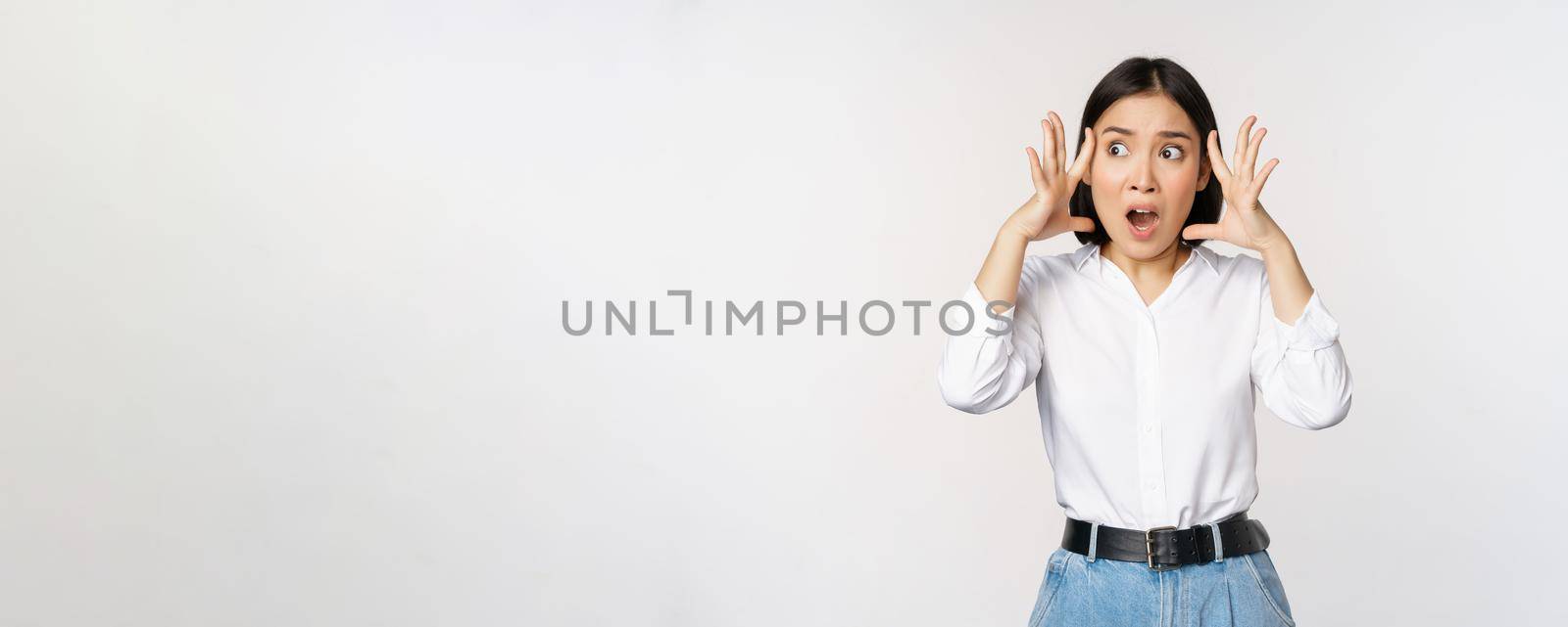 Image of shocked anxious asian woman in panic, holding hands on head and worrying, standing frustrated and scared against white background.