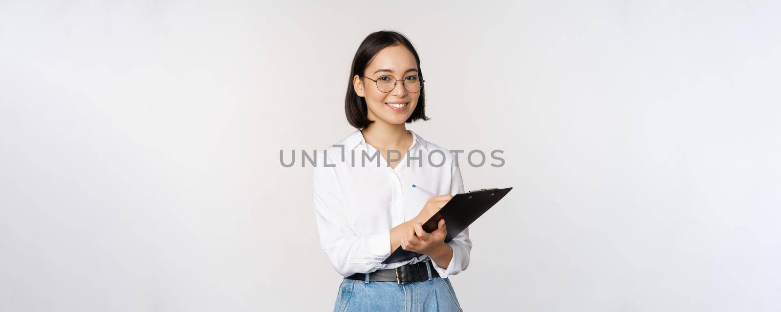 Image of smiling korean office lady, company employee writing down on clipboard, taking notes, standing over white background by Benzoix