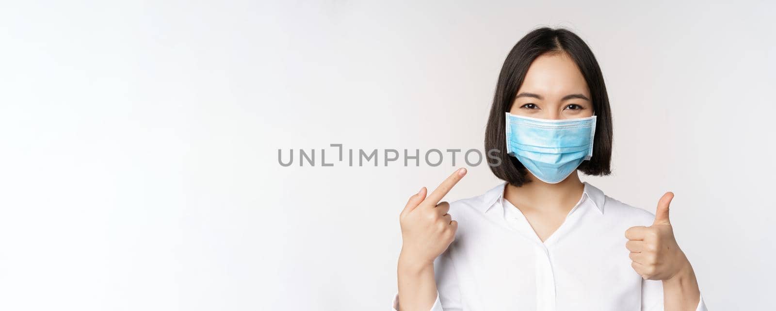 Portrait of asian girl in medical mask showing thumbs up sign and pointing at her covid protection, standing over white background by Benzoix