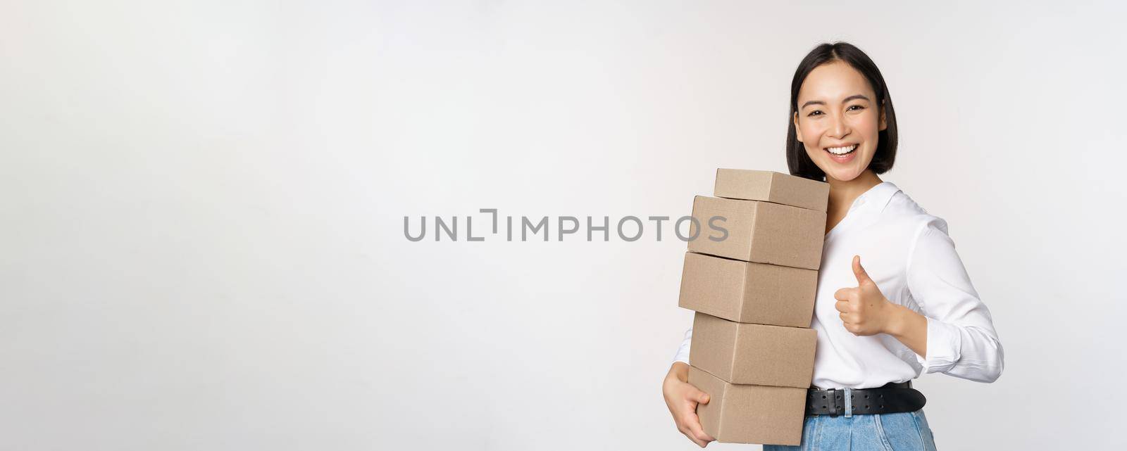Image of happy modern asian woman showing thumbs up, holding boxes delivery goods, standing against white background.