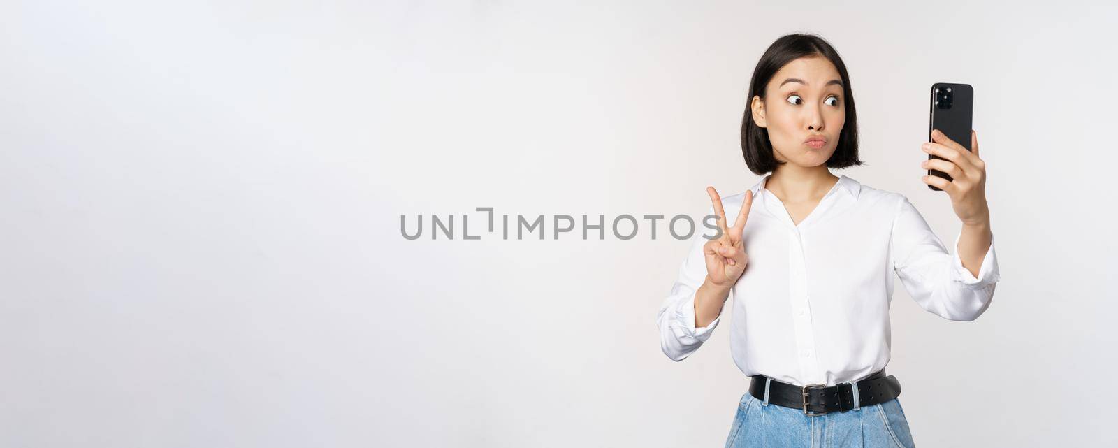 Funny asian girl showing peace, v-sign at smartphone camera, posing for photo, taking pictures with mobile application, standing over white background.