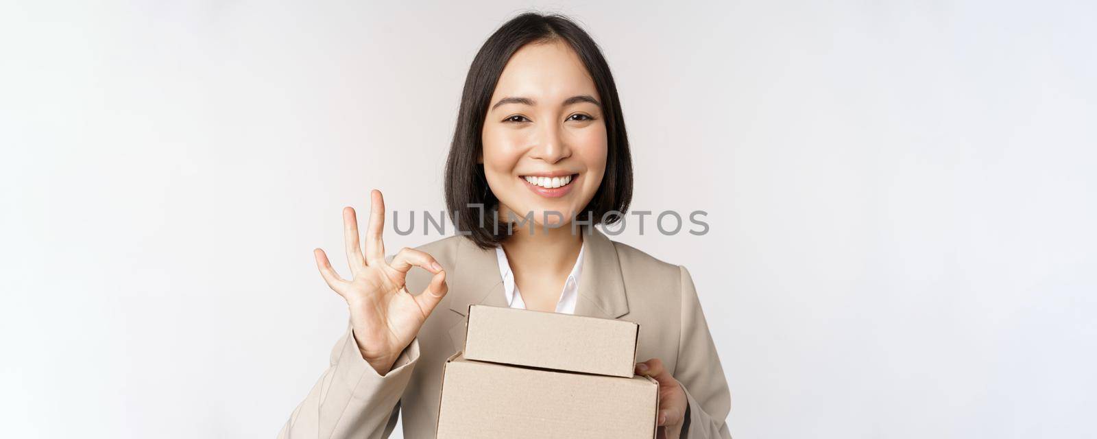 Smiling asian businesswoman, showing okay sign and boxes with delivery goods, prepare order for client, standing over white background by Benzoix