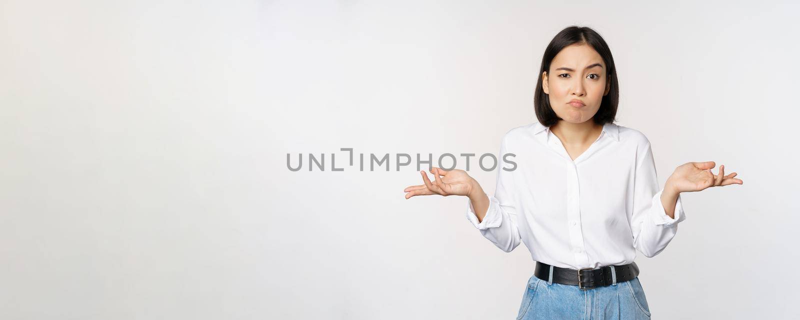 Image of confused asian office manager, office lady shrugging shoulders and looking clueless, standing puzzled against white background.