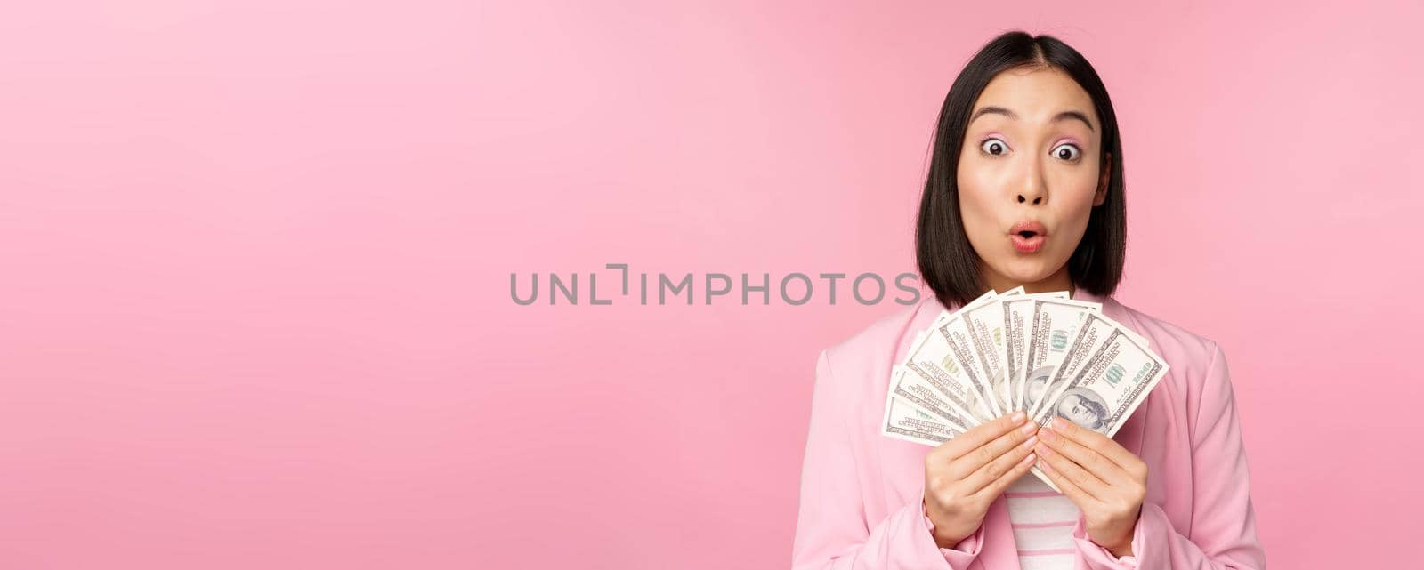 Finance, microcredit and people concept. Happy smiling asian businesswoman showing dollars money, standing in suit against pink background by Benzoix