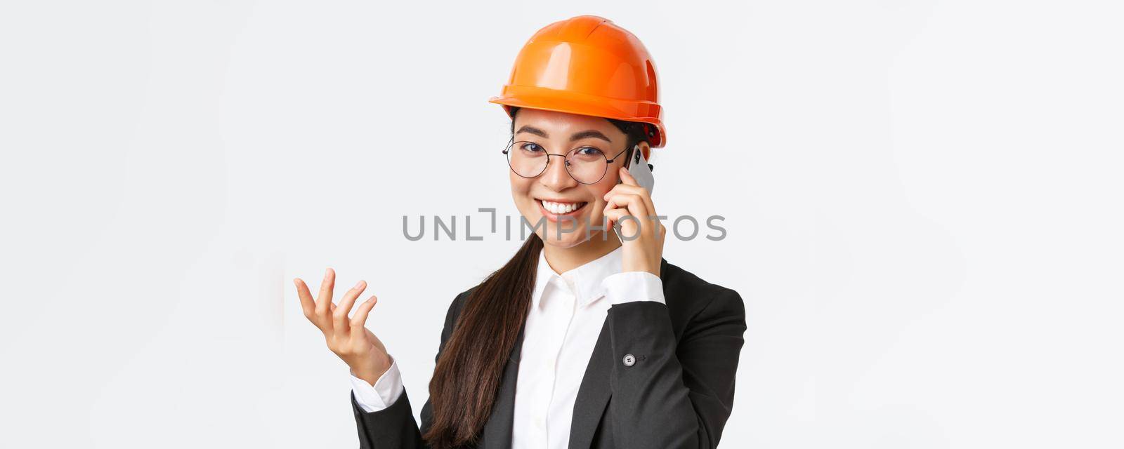 Close-up of asian businesswoman manage enterprise, engineer in safety helmet and suit having phone conversation, calling investors, smiling while talking over smartphone, white background.