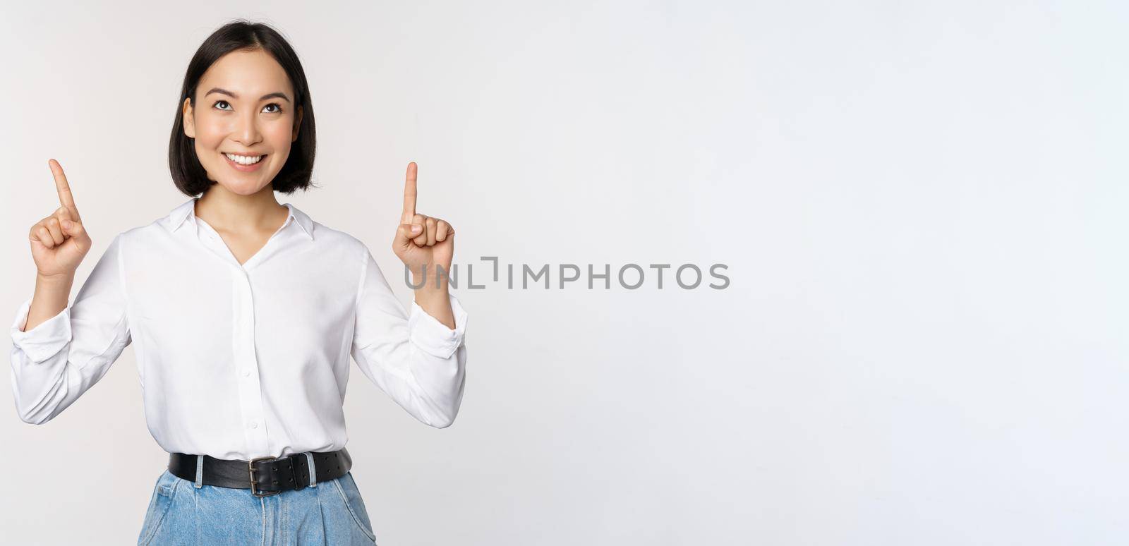 Enthusiastic asian business woman pointing, looking up with happy smiling face, showing company logo or banner, standing over white background.