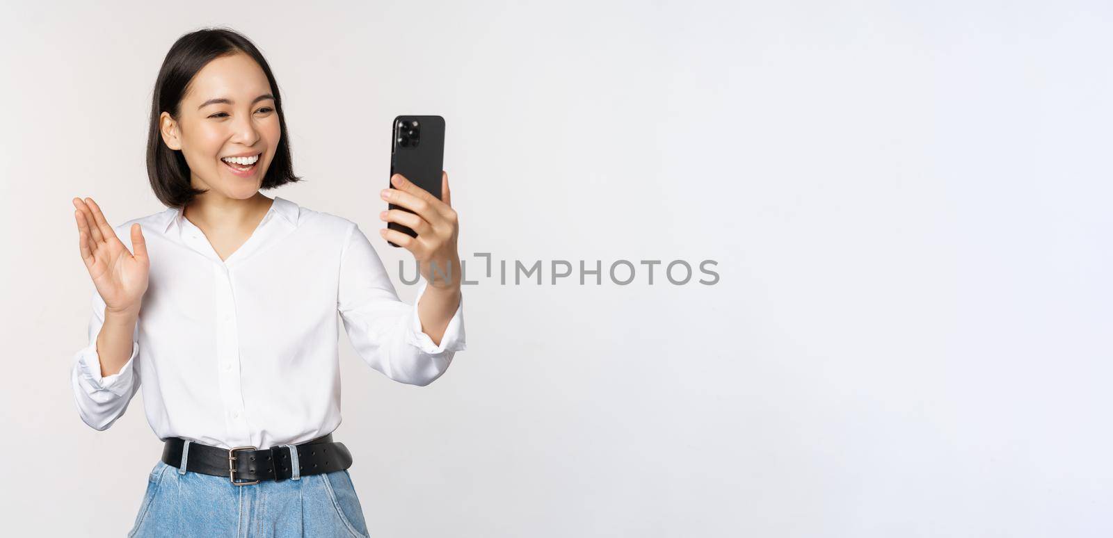 Image of stylish modern asian woman waving hand at smartphone front camera, video chat, chatting with person on mobile phone application, white background.