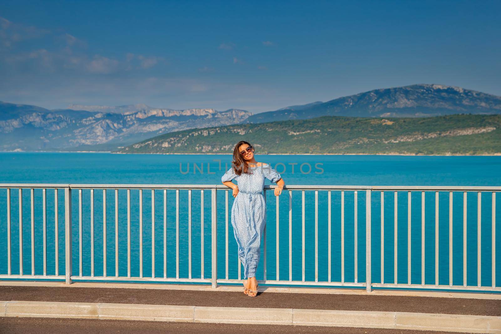 The beautiful girl in a blue dress and sunglasses poses on the bridge, the long chestnut hair, happy and smiles, azure water of the lake and slopes of mountains on a background. High quality photo