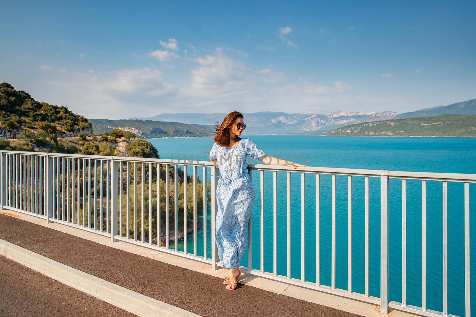 The beautiful girl in a blue dress and sunglasses poses on the bridge, the long chestnut hair, happy and smiles, azure water of the lake and slopes of mountains on a background. High quality photo