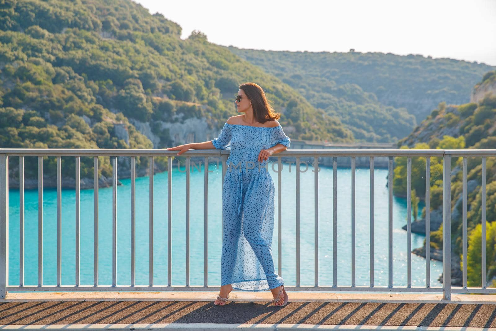 The beautiful girl in a blue dress and sunglasses poses on the bridge, the long chestnut hair, happy and smiles, azure water of the lake and slopes of mountains on a background. High quality photo