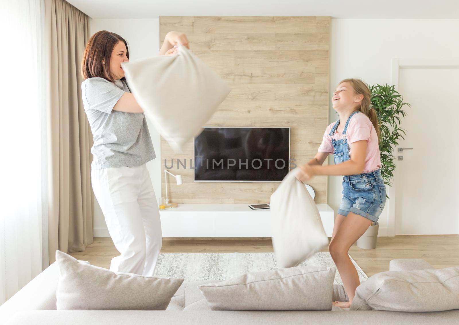 Happy family games. Single mother and her child girl are fighting pillows and jumping on couch by Mariakray