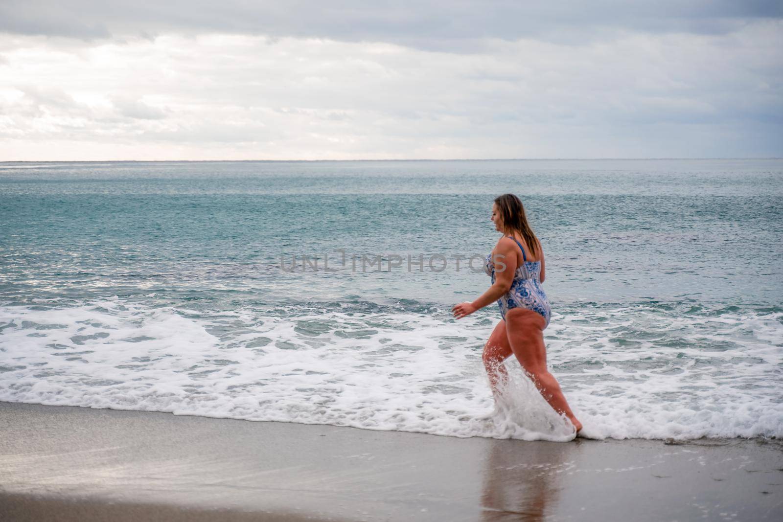 A plump woman in a bathing suit enters the water during the surf. Alone on the beach, Gray sky in the clouds, swimming in winter