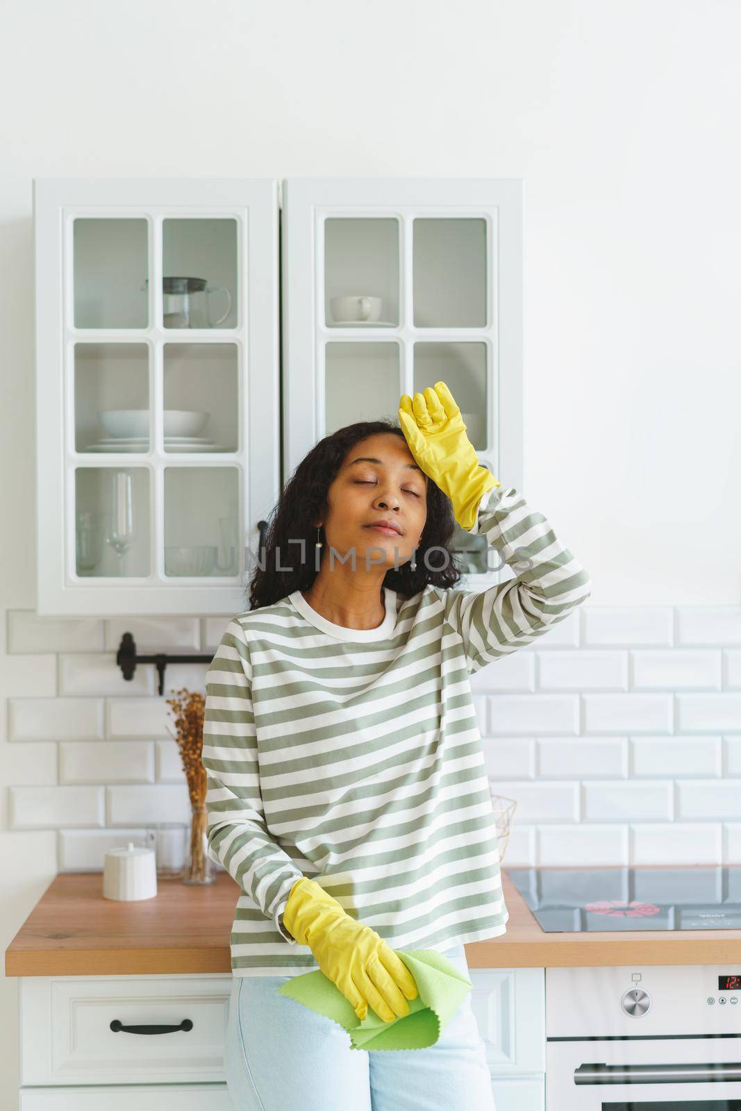 Concept of African-American woman wiping sweat from forehead after finishing doing household chores. Having rest after cleaning kitchen unit in rubber gloves with washcloth