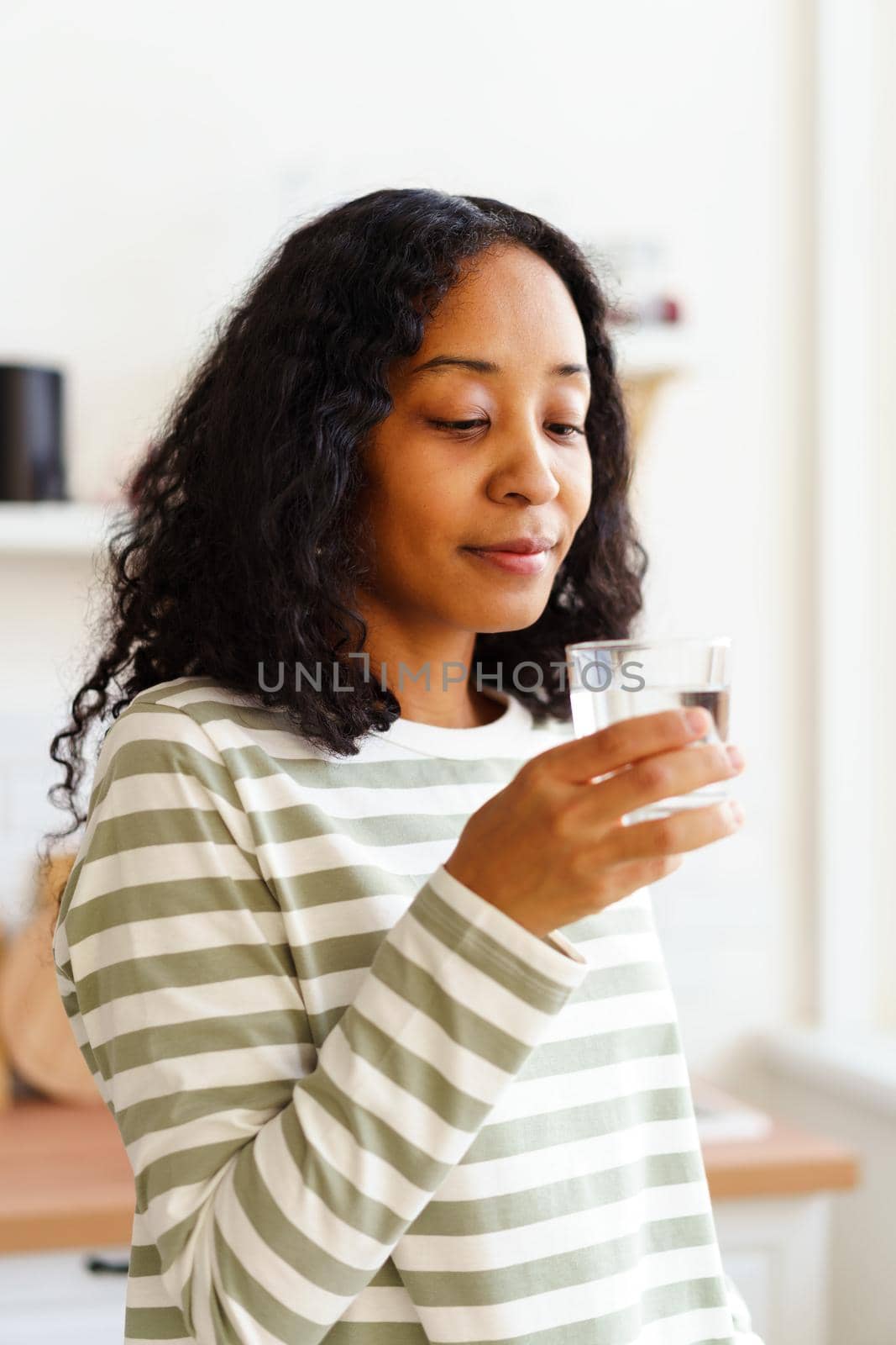 Delightful African-American woman in comfy home clothing holding glass of water. Healthy start of day, refreshing morning beginning. Concept of dieting lifestyle. Young female quenching her thirst