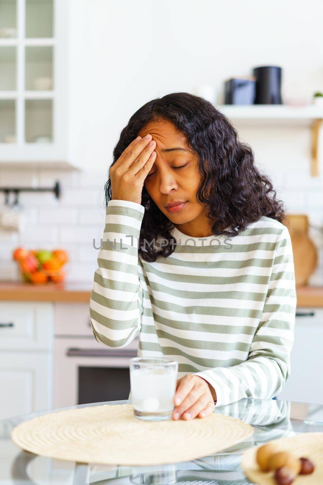 Concept of African-American woman having terrible headache. Dissolving fizz drug in glass of water in kitchen. Taking painkiller to cure pain. Touching forehead. Vertical