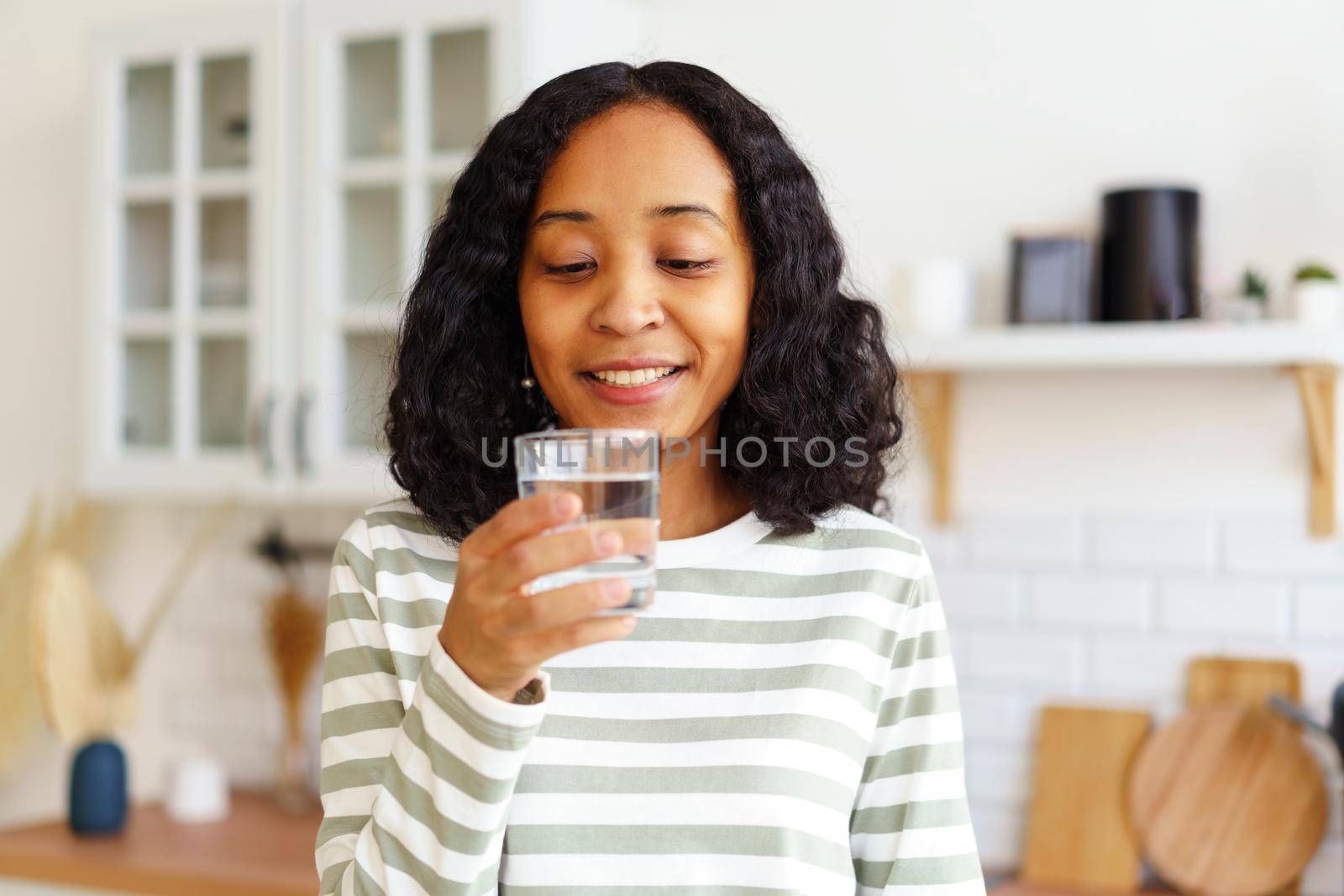 Happy and delightful young African-American woman looking at glass of clear water and ready to take sip in kitchen. Selective focus, soft lighting, horizontal, copy space