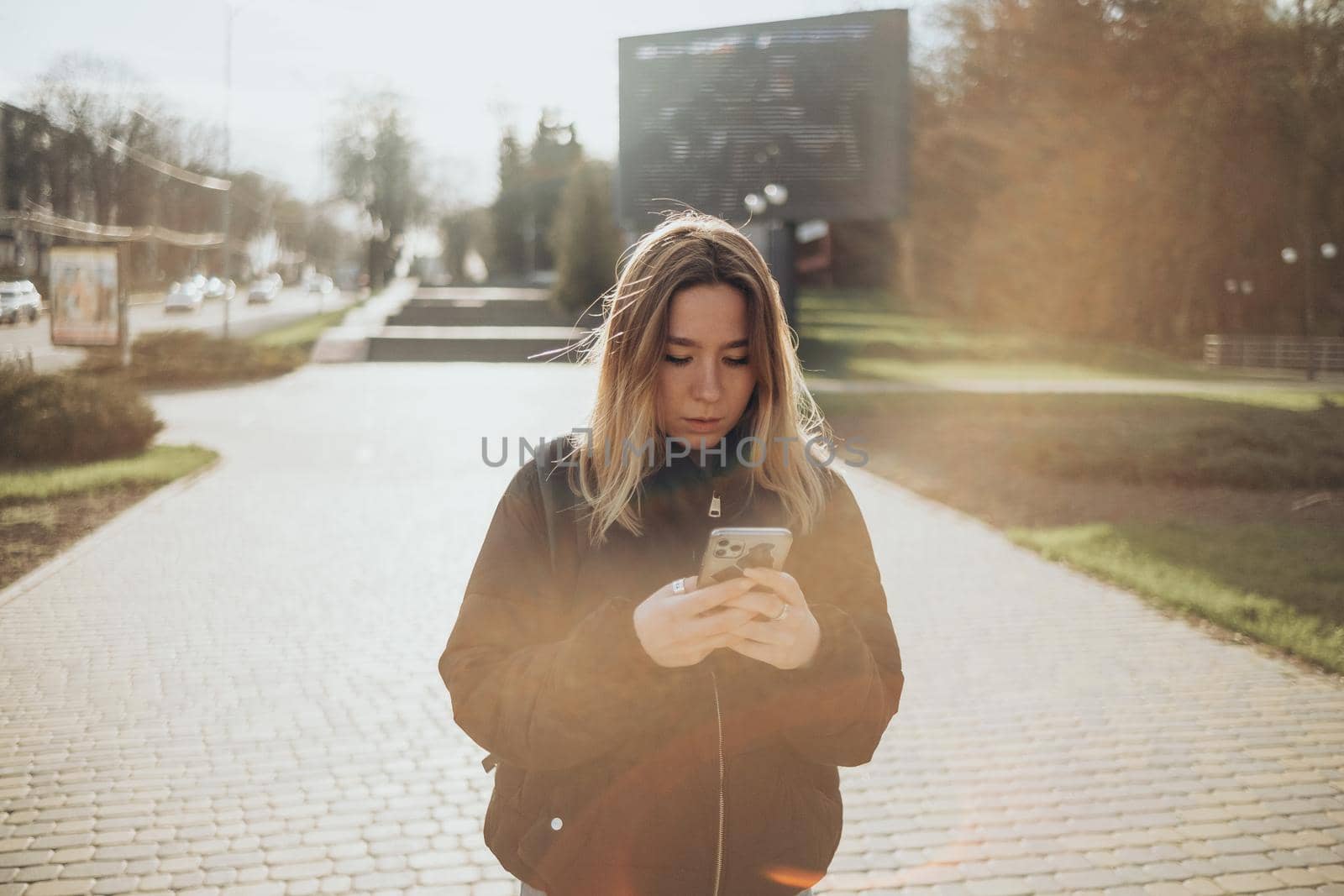 gorgeous beautiful young woman with blonde hair messaging on the smart-phone at the city street background. pretty girl having smart phone conversation in sun flare.