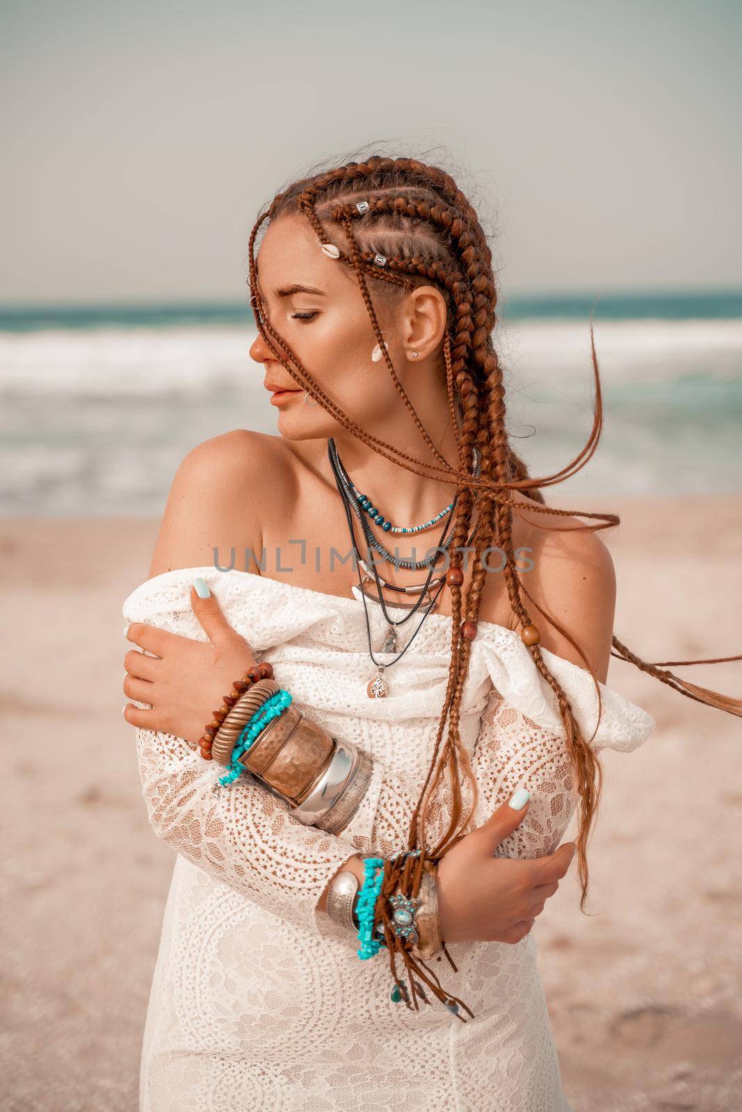 Model in boho style in a white long dress and silver jewelry on the beach. Her hair is braided, and there are many bracelets on her arms. by Matiunina