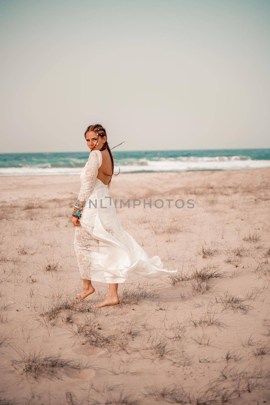 Model in boho style in a white long dress and silver jewelry on the beach. Her hair is braided, and there are many bracelets on her arms. by Matiunina