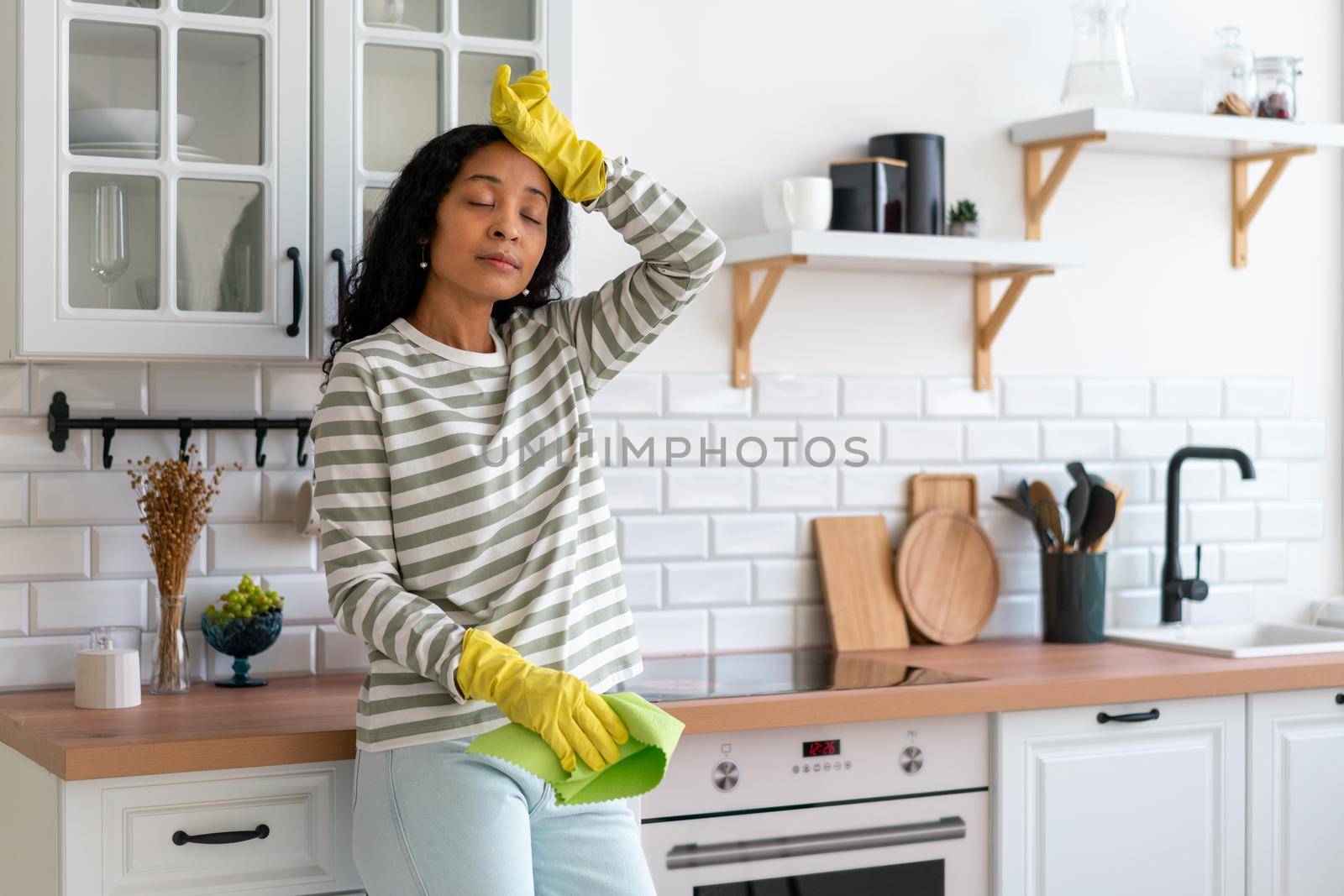 African-american woman wiping sweat from forehead after cleaning kitchen unit. Having rest after dust handling in rubber gloves with washcloth. Concept of tired housekeeper. Finishing cleanup