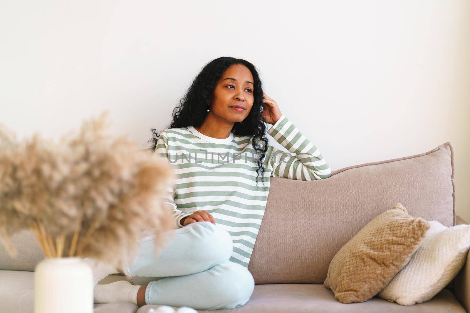 Happy young african-american female sitting on couch in living room. Concept of enjoying life and slow pace lifestyle. Thoughtful smiling woman self-reflecting
