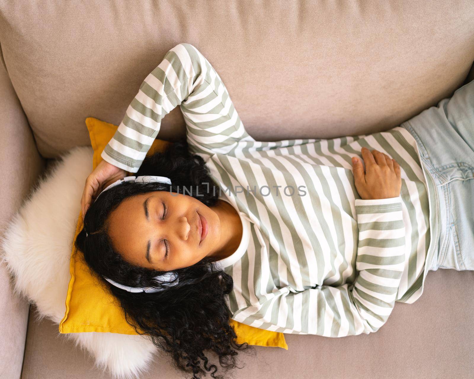 Happy african-american woman lying on sofa and listening to music in headphones. Enjoying audio playing in earphones. Relaxing and chilling at home. Positive emotions. Having fun while laying along