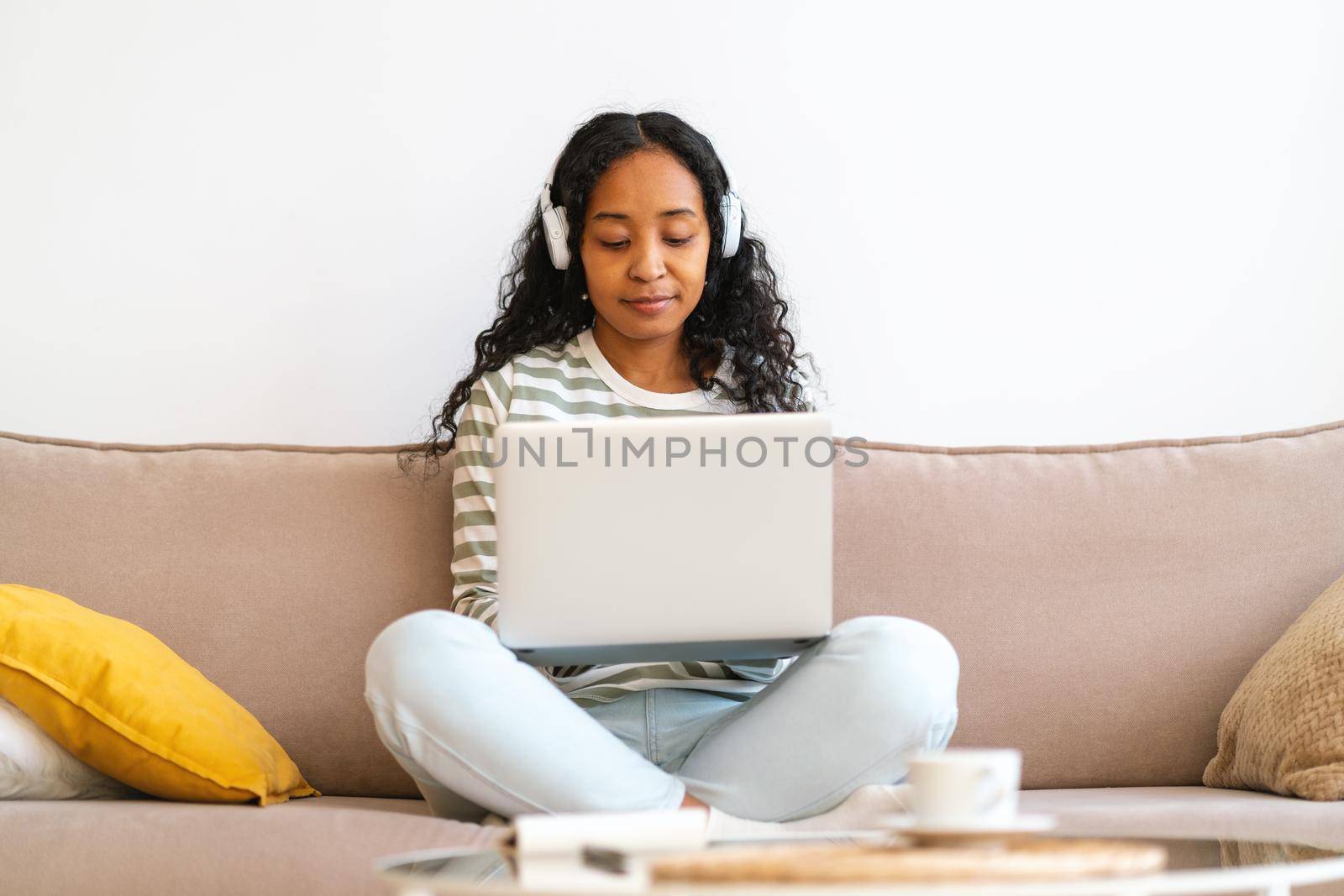 Concentrated african-american female listening to online lecture in headphones while searching information on laptop. Sitting on sofa and studying from home. Advantages of e-learning