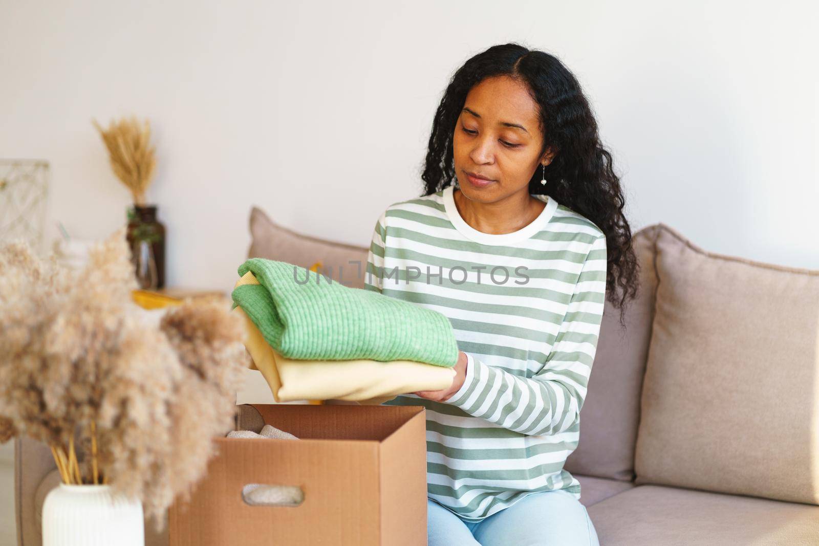 Young african-american female in casual packing clothes for charity donation in cardboard box while sitting on sofa in living room. Concept of shipping stuff due to move. Giving things away